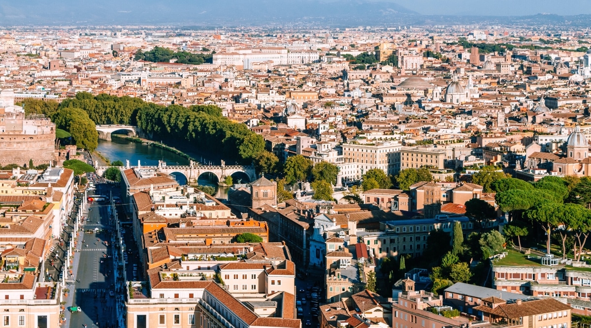 An aerial photograph capturing the cityscape of Rome. The image presents a comprehensive view of the urban layout, featuring streets, buildings, and landmarks. The Tiber River winds through the city.