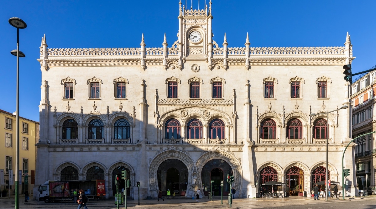 A photograph capturing Rossio Railway Station in Lisbon. The image features the station's architectural facade and surrounding urban environment. Notable elements include the station's arched entrance, clock tower, and a few pedestrians in the vicinity.