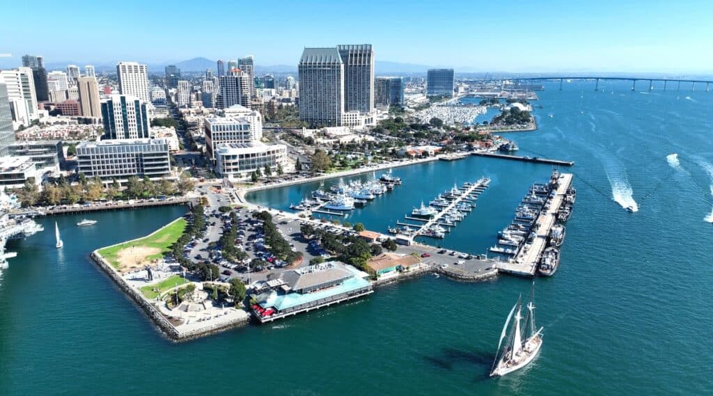 A panoramic view capturing the coastal area of San Diego, California. The image showcases a pier with moored yachts and boats along the waterfront. In the background, city streets are visible, flanked by tall buildings that contribute to the urban skyline. The scene depicts limited greenery, with emphasis on the built environment and coastal infrastructure.