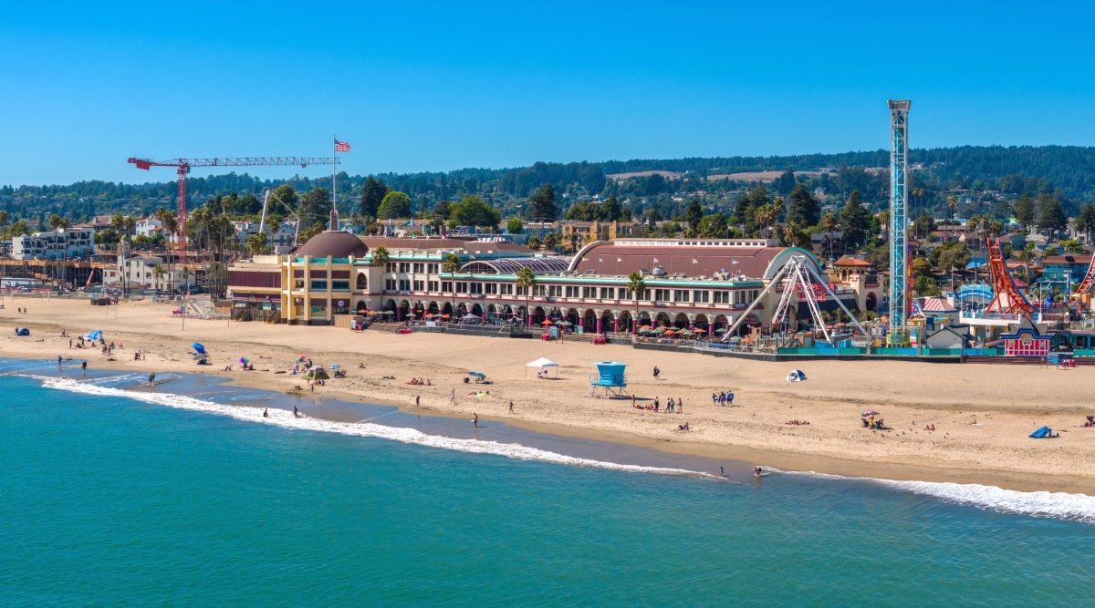 A direct view of Santa Cruz Beach with individuals engaging in swimming and relaxation along the shoreline. The background features hotels, attractions, and abundant greenery, contributing to the overall beachscape.