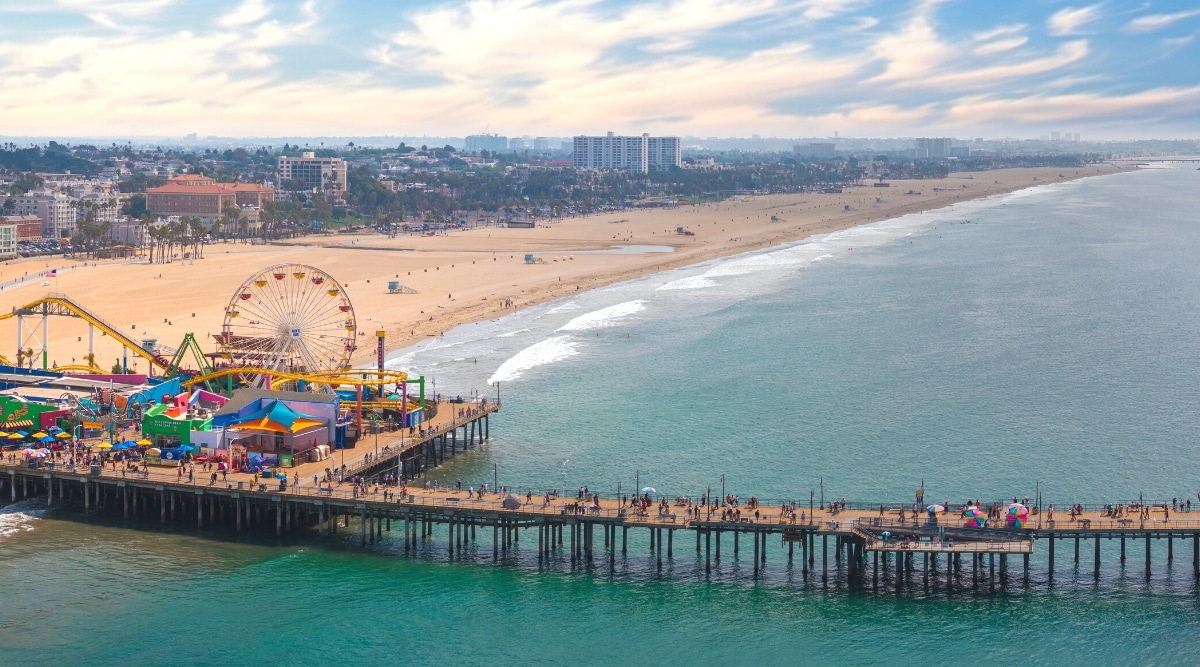 A top view capturing the expansive width of Santa Monica Beach in California. The image depicts the beach extending on the left, bordered by the cityscape, while the Pacific Ocean spans the right side. The beach is notably wide, accommodating various recreational activities. On the bottom left, a pier with attractions and people walking can be observed.