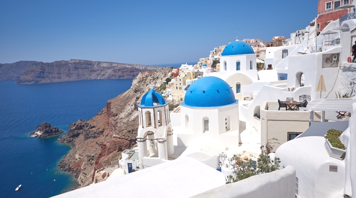 A photograph depicting the scenic landscape of Santorini, Greece. The image captures the island's distinct architecture, characterized by white-washed buildings with blue-domed roofs, against a backdrop of the Aegean Sea. 