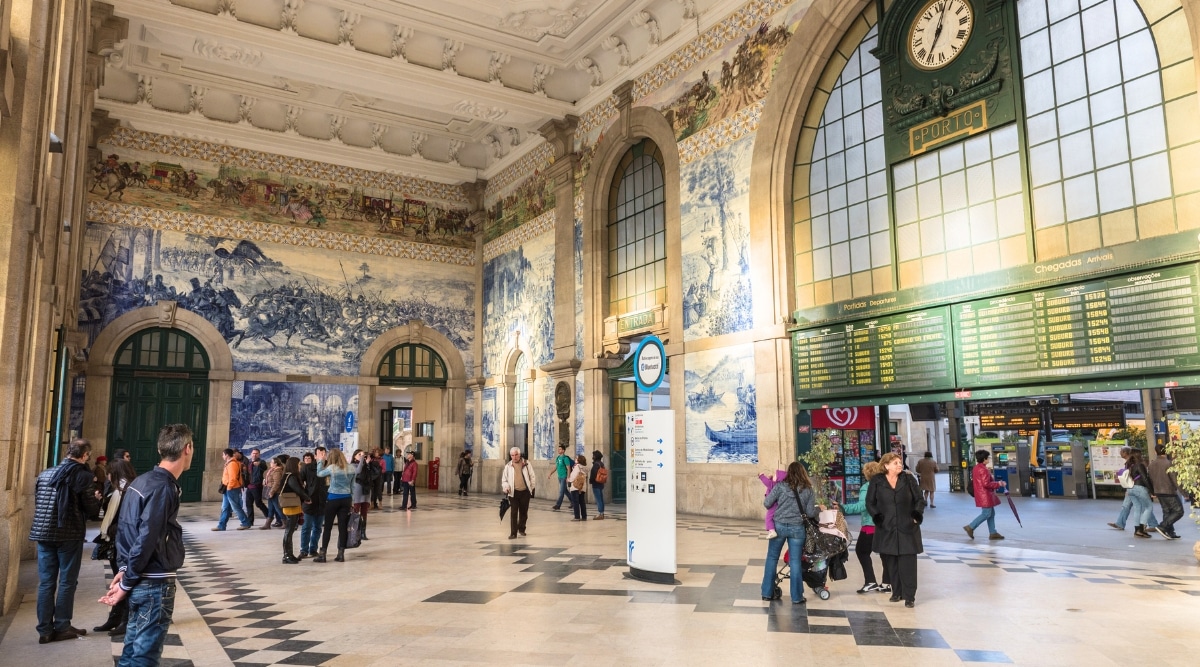 A photograph capturing the interior of Sao Bento Train Station in Porto. The image showcases the station's architectural elements, including ornate tilework and structural features. Commuters and travelers can be observed within the station, engaged in various activities such as waiting, walking, or navigating the space.