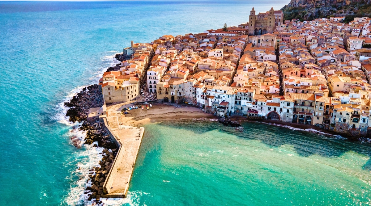 A top view capturing a cluster of densely packed brown-roofed buildings in Sicily, Italy. The photograph reveals an urban landscape characterized by compact architecture. The calm sea provides a backdrop to the residential structures.