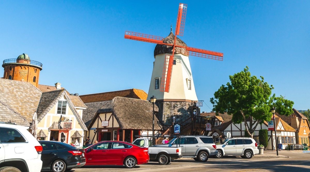 A city street in Solvang, featuring Danish-style architecture with a view of a mill. Cars are neatly parked along the street, contributing to the urban landscape. The image captures the distinctive design elements characteristic of the Danish-inspired buildings in Solvang.