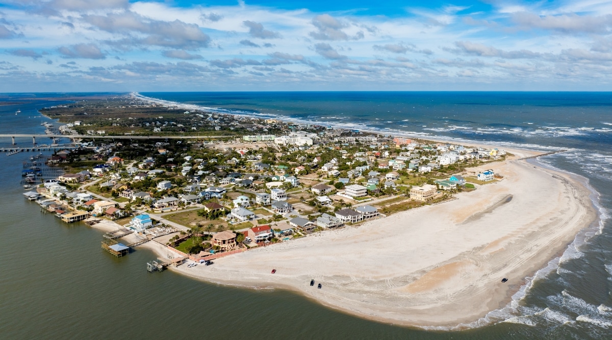 A view of St. Augustine, Florida, captured from an elevated perspective. In the foreground is a spacious beach, devoid of people. The beach extends to meet the shoreline in a calm and serene manner. Beyond the beach, a low-rise town emerges, contributing to the coastal landscape.