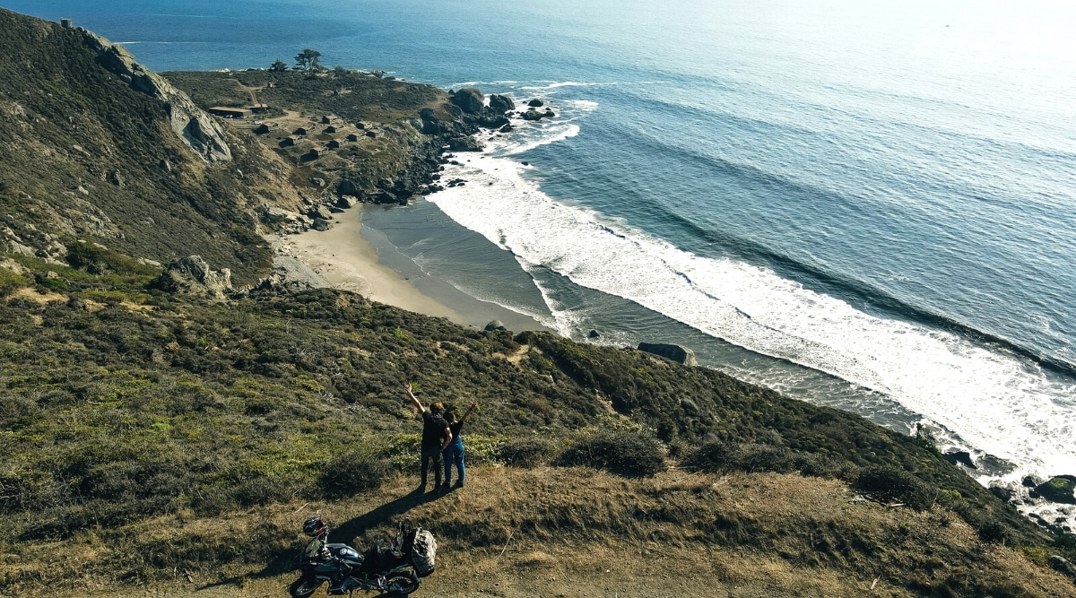 An aerial view capturing Stinson Beach in Marin County. The image features several people observing the beach, with a motorcycle present nearby. The ocean exhibits small waves.