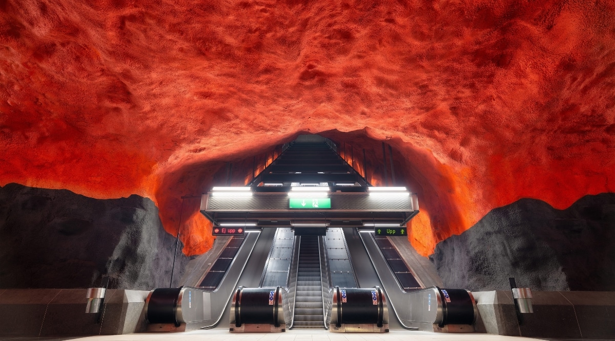 The image depicts the up and down escalators within Stockholm Metro Stations, Sweden. The escalators are situated in a minimalist environment with a rocky red ceiling. 