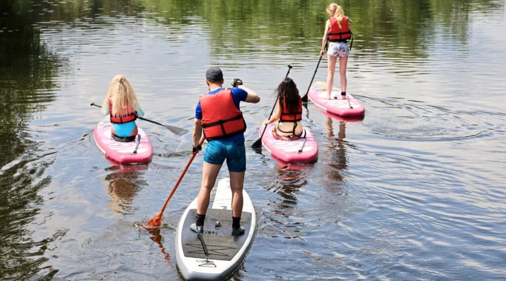 The image depicts stand-up paddle boarding (SUP) on a river during summer. A man and three girls navigate the water on individual paddle boards, engaging in SUP surfing.