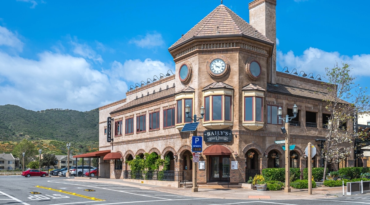 The photograph captures a streetscape in Temecula's Old Town neighborhood. The focal point is the beige stone structure housing Bailys restaurant and nightclub situated on a street corner. A backdrop of a blue cloudscape and distant mountains provides context to the local geography.