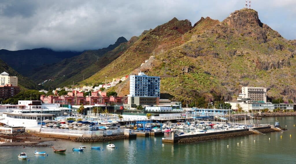 A view of Tenerife showcasing numerous boats and motorboats in the foreground, followed by city houses and, in the background, towering mountains. The sky is characterized by dark cloud cover, contributing to an overcast atmosphere.