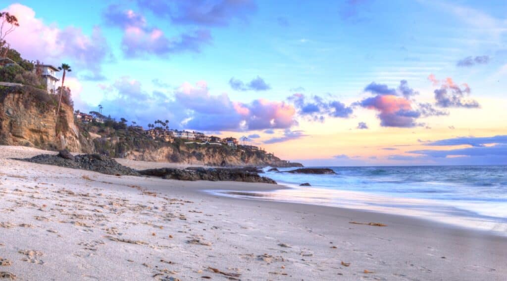 The image depicts a view of Thousand Steps Beach in Laguna. The ocean is visible on the right, and on the left is the beach with small rocks, adorned with palm trees and houses. The sky features beautiful pink clouds, contributing to the overall atmospheric conditions.