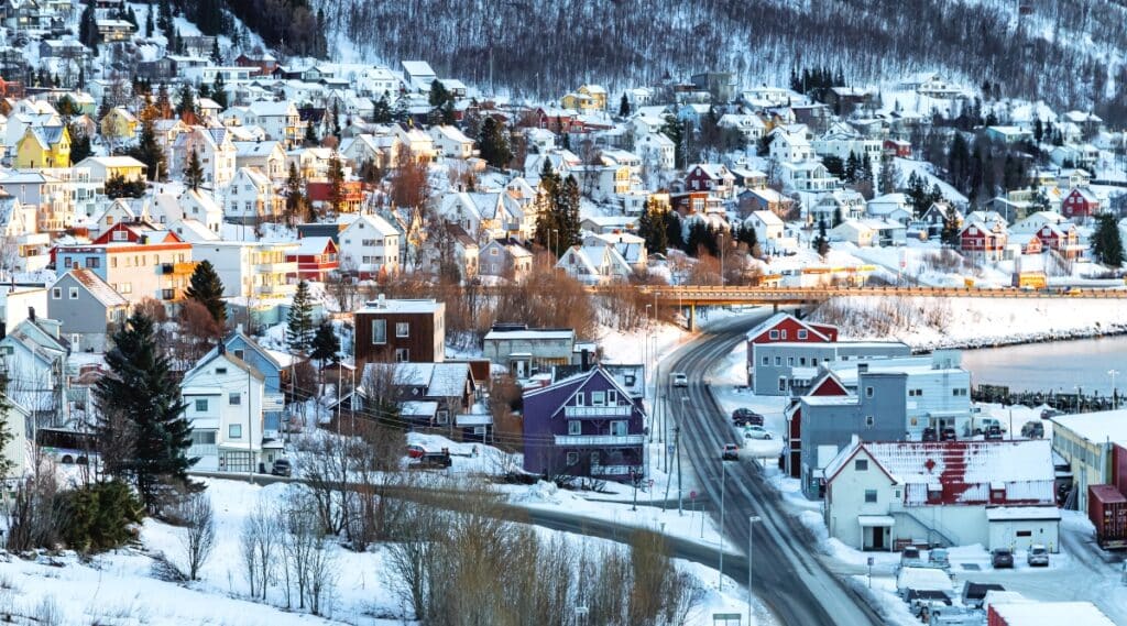A winter view capturing a very snowy landscape in Tromsø, Norway. The image features many bright houses nestled in the snow-covered terrain, surrounded by numerous trees. 