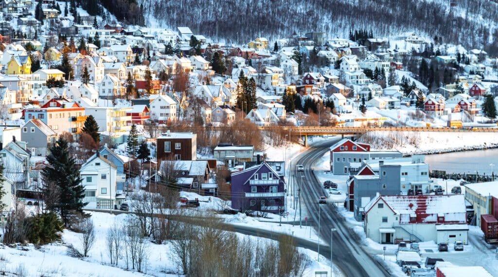 A winter view capturing a very snowy landscape in Tromsø, Norway. The image features many bright houses nestled in the snow-covered terrain, surrounded by numerous trees.