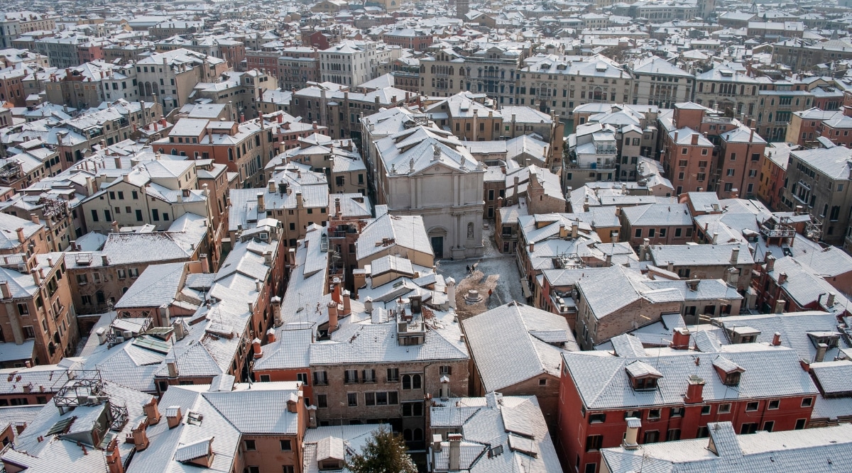 A top-view photograph depicting a snow-covered Venice, Italy. The image captures the snowy rooftops of the city's buildings, presenting a serene winter scene.
