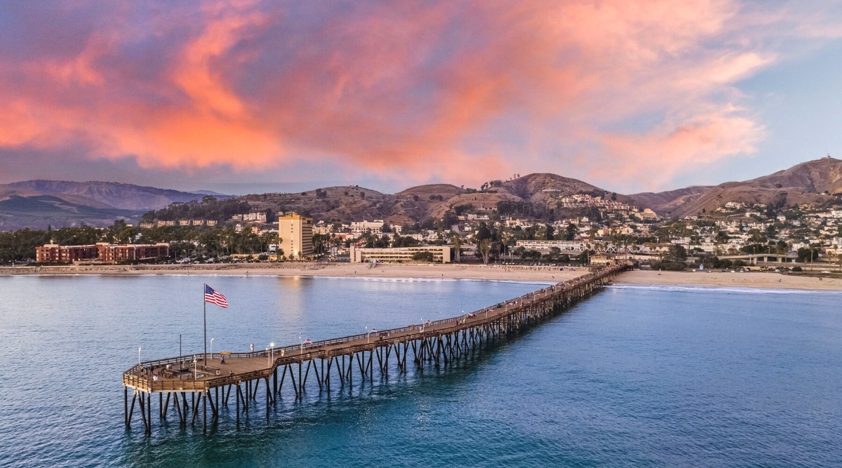 An image capturing a sunset scene over Ventura Beach, California, with a long pier extending into the ocean. The photograph features a vibrant sky with warm hues, reflecting the concluding moments of the day.