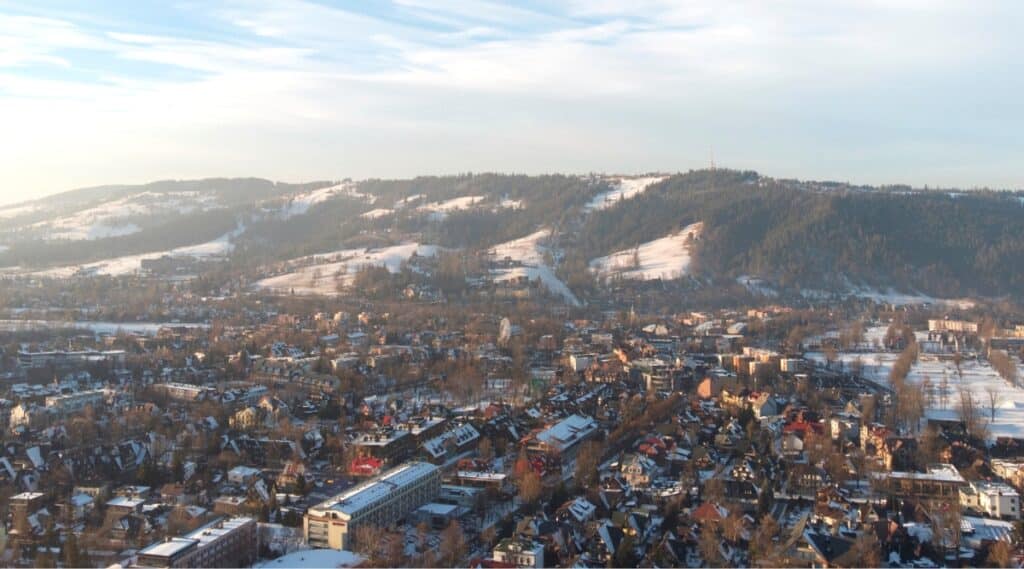 A drone view capturing the snowy cityscape of Zakopane, Poland. In the foreground, the city is depicted with numerous trees, while in the background, a long snow-covered hill extends across the scene. The aerial perspective provides a comprehensive overview of the winter landscape in Zakopane, emphasizing the snow-covered urban and natural elements.