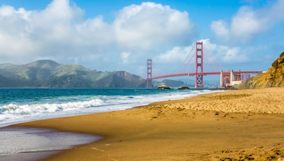 A direct view of Baker Beach in San Francisco, with the coastline visible to the right. The image captures an absence of people. In the distance, a prominent bridge is directly visible. The weather conditions appear neutral.