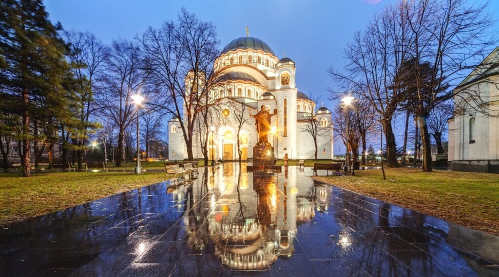 A photograph capturing the Saint Sava Temple in Belgrade during a winter night devoid of snow. The image features the iconic temple with illuminated pillars, casting light in the surrounding area. The urban setting is notably quiet, with an absence of pedestrians on the streets.The winter night ambiance is conveyed without snow, and the illuminated pillars of the Saint Sava Temple provide a focal point.