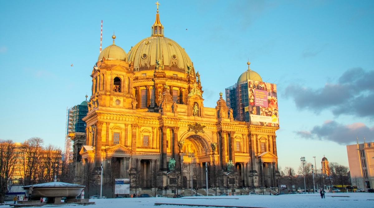 A landscape photograph capturing Berliner Dom Cathedral on a snowy day. The image showcases the iconic cathedral against a backdrop of winter weather conditions, with snow covering the surrounding landscape. The architectural details of the cathedral, including its domes and spires, are visible amidst the wintry scene.