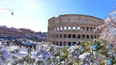The Colosseum situated in Rome, Italy, depicted on a white winter day. The surroundings are covered in a layer of snow, extending to the trees in the vicinity. Numerous visitors are observed around the Colosseum.