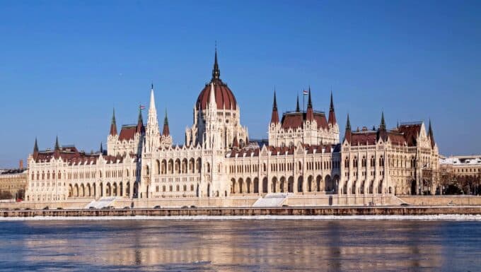 The Hungarian Parliament building in Budapest situated along the Danube River. The image provides a straightforward view of the iconic architectural structure against the cityscape. The distinctive features of the parliament, including its domes and spires, are visible in the image. The Danube River flows adjacent to the building.