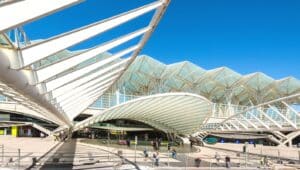 An image capturing Gare do Oriente in Lisbon, Portugal, from a distant viewpoint. The photograph depicts the modern architectural design of the transportation hub, featuring a complex structure with distinctive geometric patterns. Several people are observed in the vicinity.