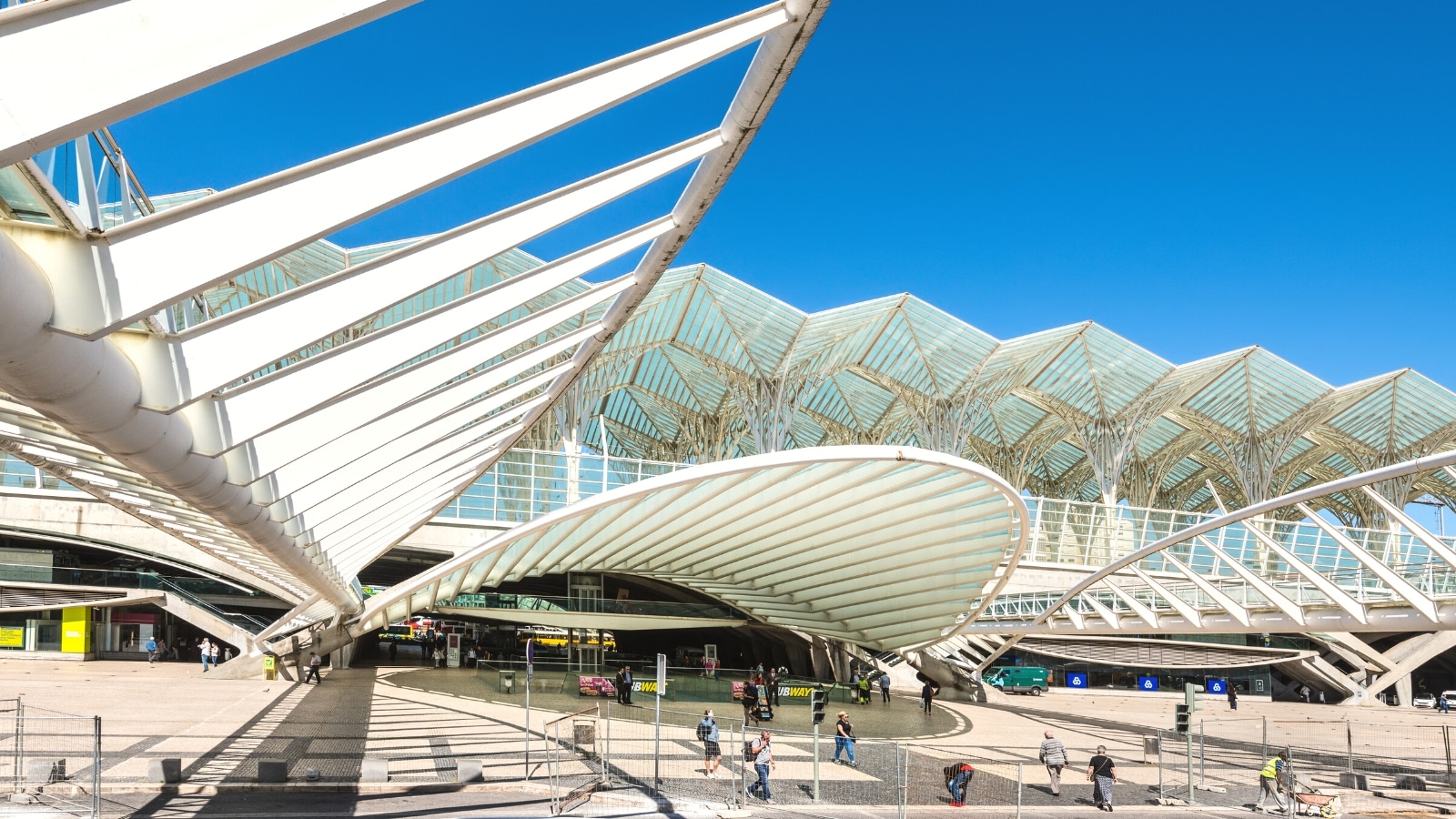 An image capturing Gare do Oriente in Lisbon, Portugal, from a distant viewpoint. The photograph depicts the modern architectural design of the transportation hub, featuring a complex structure with distinctive geometric patterns. Several people are observed in the vicinity.