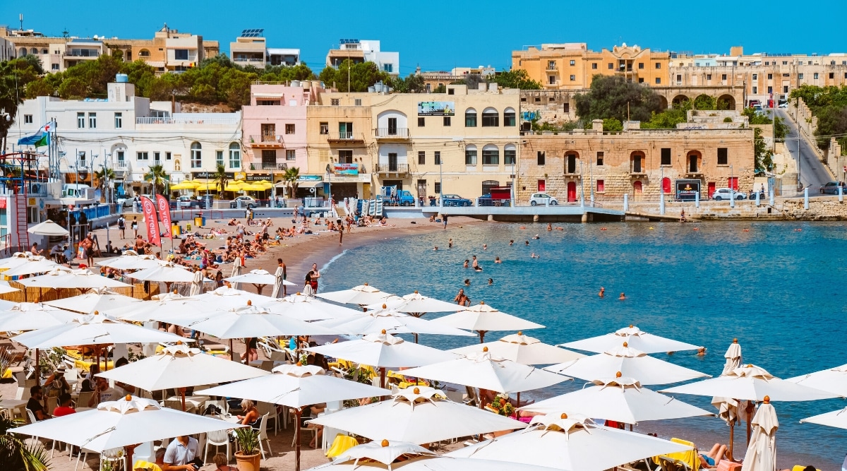 An aerial view capturing a beach scene in Malta, featuring a backdrop of 2-3 storey buildings. The beach is adorned with large white umbrellas in the foreground. People can be seen swimming in the sea, contributing to the typical coastal activities.