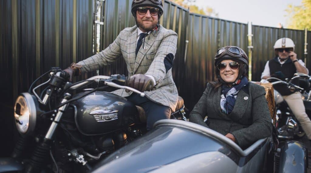 A photograph featuring a man at the wheel of a motorcycle with a sidecar, accompanied by a woman seated in the sidecar. Both individuals are attired in suits, shirts, and ties. The image captures a simple scene against a backdrop of a metal fence in the background.