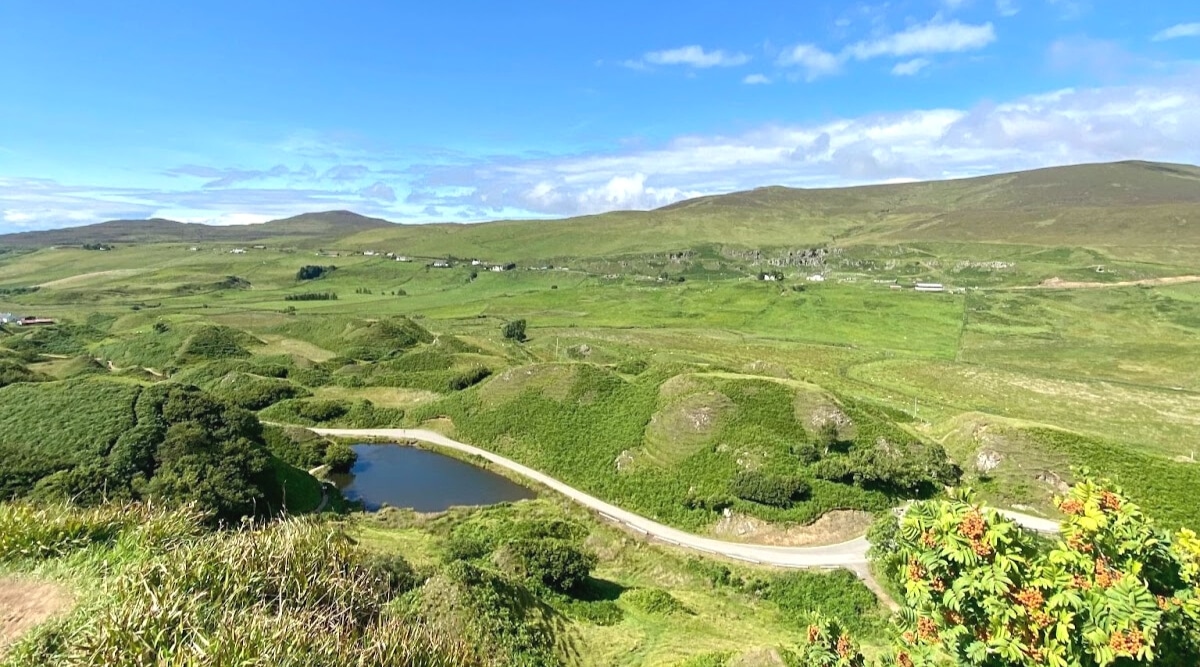 A serene natural landscape captured in Scotland, featuring hills and abundant greenery. The image includes a tranquil lake surrounded by lush vegetation, with clear paths visible throughout the scene. The sky is clear with a few scattered clouds. 