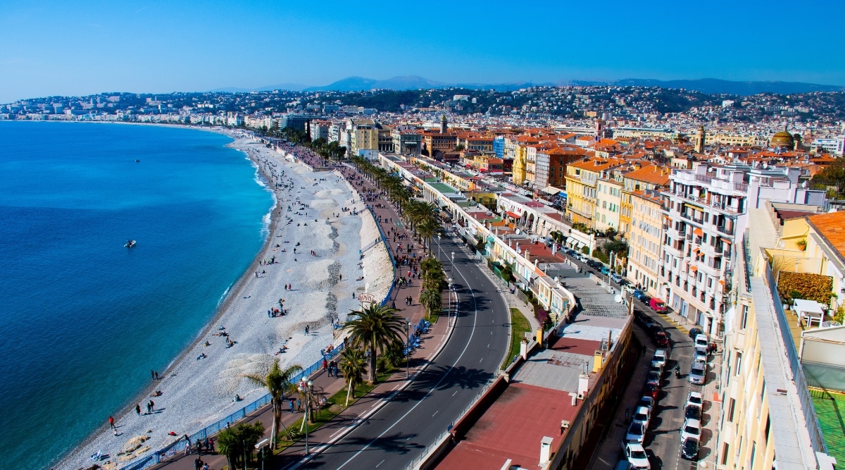 An aerial drone view capturing the coastline of Nice, located on the French Riviera, on a sunny day. The image provides an expansive overview of the coastal landscape, extending from the azure Mediterranean waters to the city's architectural structures.