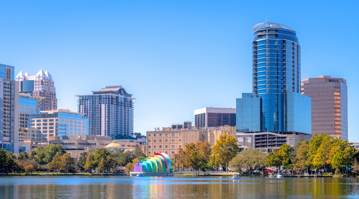 A direct view of Orlando, Florida, featuring a river in the foreground and a tall cityscape in the background. Along the riverbank, trees line the landscape. The city skyline exhibits architectural prominence with clear, unobstructed views.