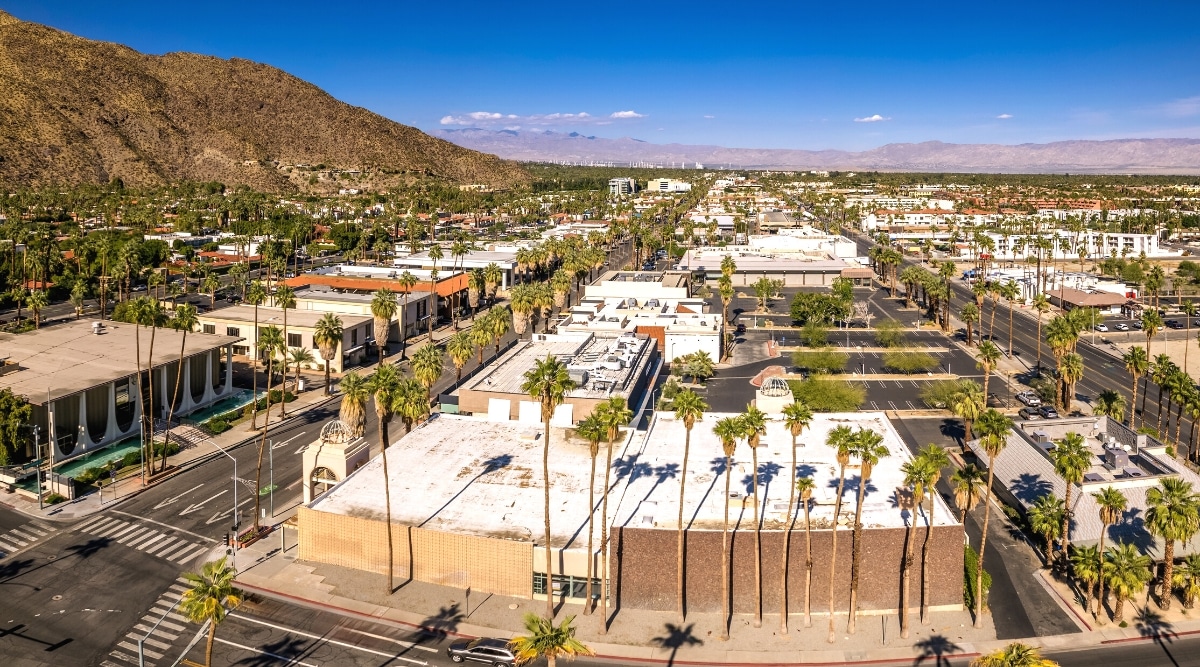 A top view capturing Palm Springs, California, on a bright day with a clear sky. The image features palm trees, white houses, a few cars, and people, creating a typical suburban scene. Hills are visible on the left side of the image, contributing to the overall landscape.