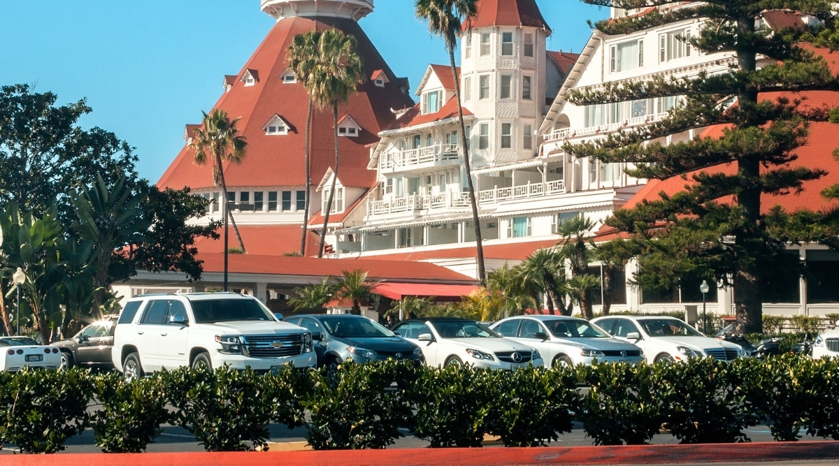 A photograph capturing the parking area adjacent to Hotel Del Coronado, featuring cars of various brands. The parking lot is surrounded by ample greenery. The cars are neatly parked. In the background is Hotel Del Coronado.