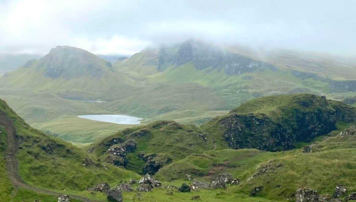 The image depicts a mountain valley in Scotland characterized by hills adorned with green vegetation. A small lake with clear water is visible within the valley. The mountain summits are capped with thick white mist.