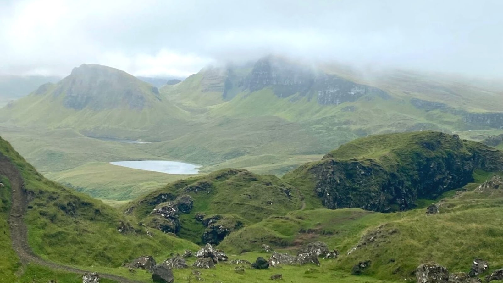 The image depicts a mountain valley in Scotland characterized by hills adorned with green vegetation. A small lake with clear water is visible within the valley. The mountain summits are capped with thick white mist.