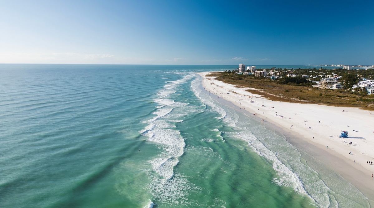 A top-down view capturing Siesta Key, Florida, with a clear sky, the ocean on the left, and a beach and city buildings on the right. Tall multi-story buildings are visible, contributing to the coastal cityscape. The beach is populated with numerous individuals engaged in recreational activities.