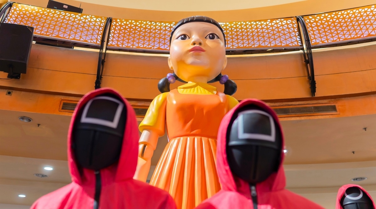 A photograph featuring characters from the Squid Game series in red overalls standing next to a doll decoration in an orange dress in Los Angeles. The image captures the characters in a staged setting, replicating scenes from the popular series.