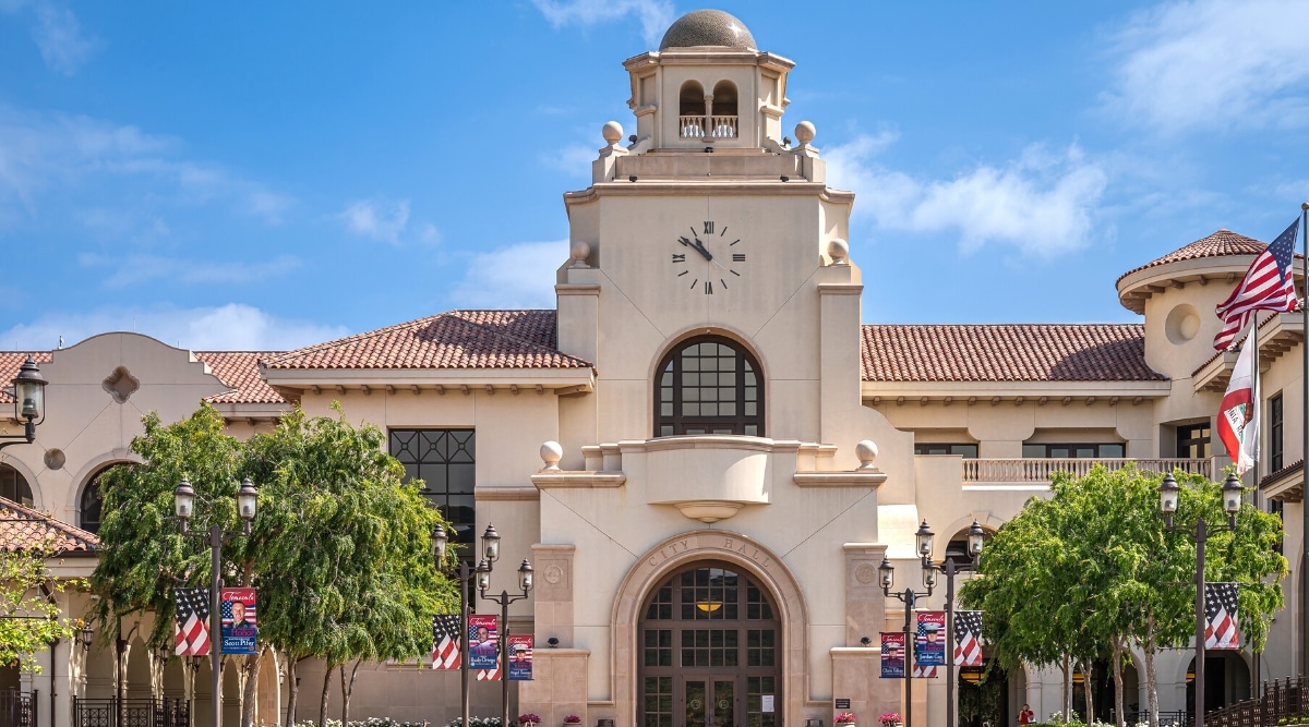 A close-up depiction of the town hall building in Temecula's Old Town neighborhood, featuring a clock tower under a blue cloudscape. The image includes a row of USA flags. Architectural details of the town hall, including the clock tower, are visible in this straightforward representation.