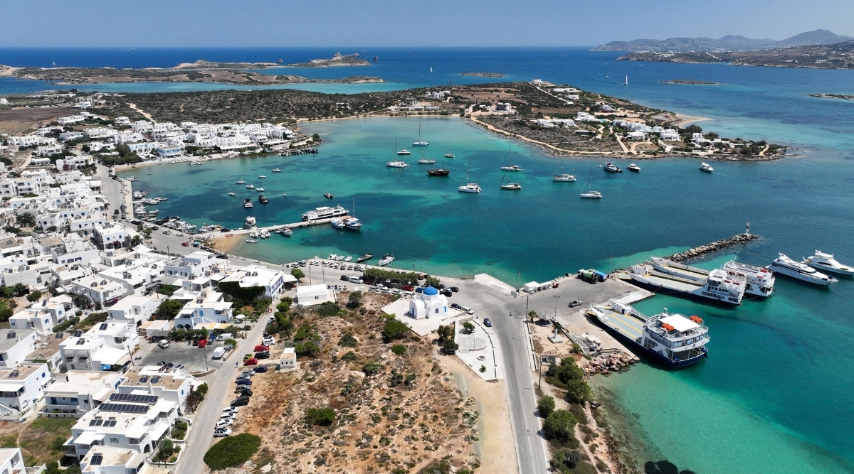 An aerial drone photograph capturing the picturesque port and main village, Hora, of Antiparos island in the Cyclades, Greece. The image offers a comprehensive view of the coastal area, showcasing the architectural layout of the village against the backdrop of the Aegean Sea. Notable landmarks and structures are visible, providing a glimpse into the island's urban landscape.