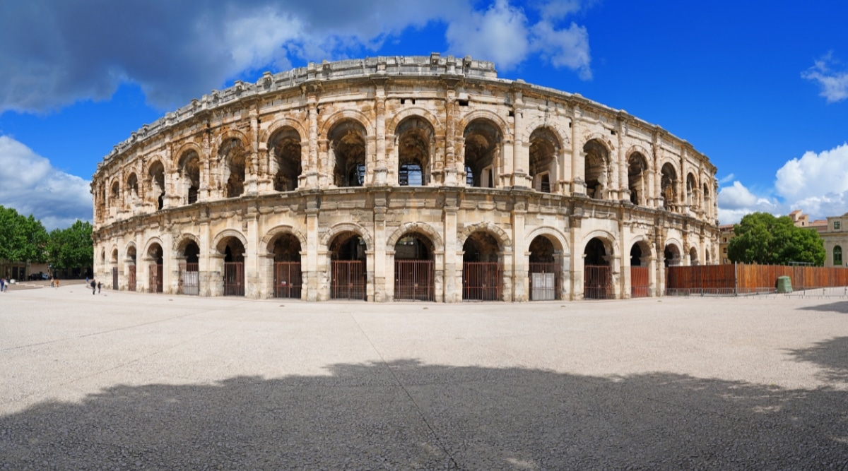 A photograph capturing Arenes de Nimes in Nimes, France. The image provides a straightforward view of the amphitheater's exterior, showcasing its historical architecture and scale. The arena stands against a clear sky, surrounded by urban structures.