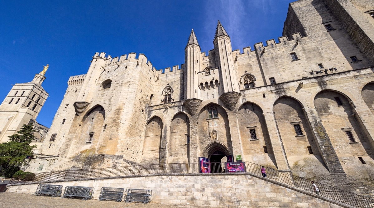 A photograph capturing the cityscape of Avignon, featuring its ancient streets, a restored medieval fortress, and the monumental Gothic architecture of the Papal Palace.