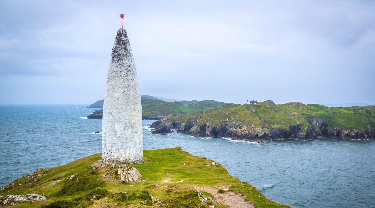 A photograph depicting Baltimore Beacon in County Cork, Ireland. The image captures the beacon against a backdrop of the Irish landscape. The beacon, a simple stone structure, stands prominently on the coastal terrain.