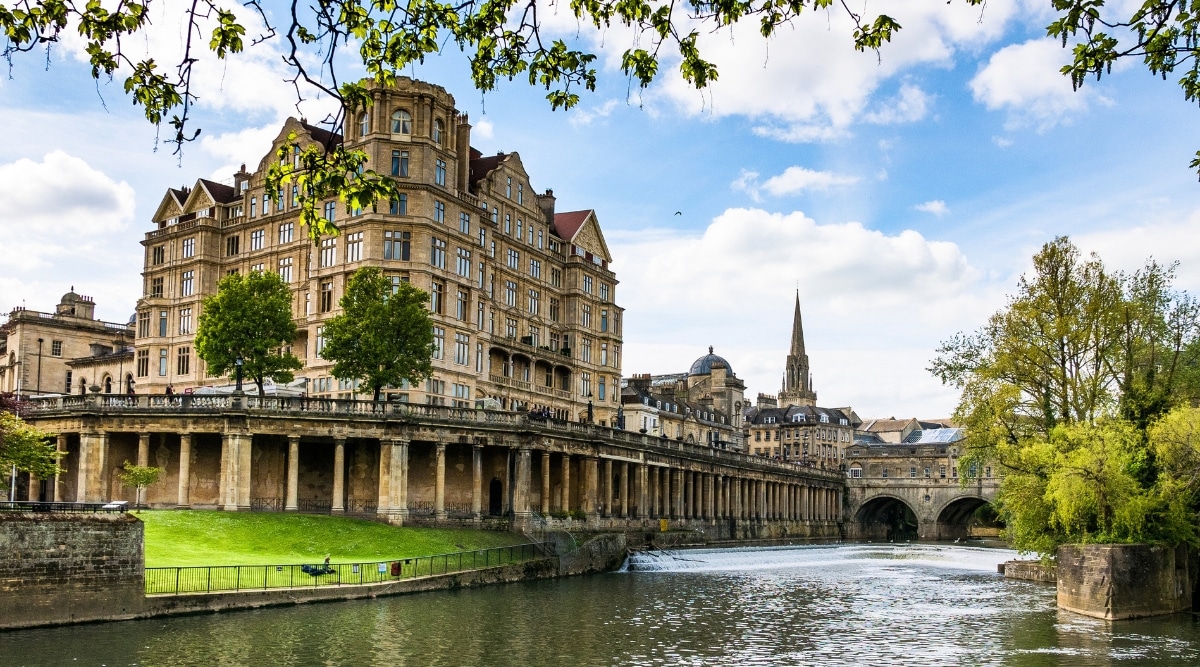 A photograph capturing a view of Bath, England. In the foreground, a river flows under a bridge, and in the background, a large, historic building adds architectural prominence to the scene. The surroundings are characterized by lush greenery.