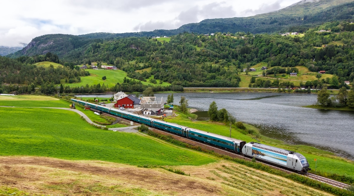 A view of the Bergen Railway in Norway featuring a train moving through a scenic natural landscape. The train travels amidst mountains, rivers, and forests, with an abundance of lush greenery. The overcast sky provides a neutral backdrop to the picturesque surroundings.