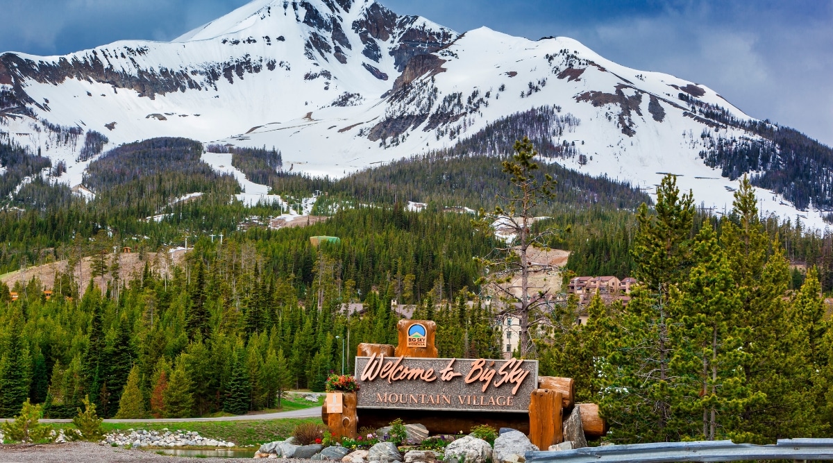 A photograph capturing a prominent sign with the inscription 'Welcome to Big Sky Mountain Village' displayed throughout the city of Big Sky, Montana. The image features a straightforward view of the sign against the cityscape. In the background, snow-capped mountains form a scenic backdrop.