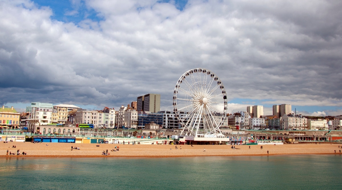 A photograph capturing the view of Brighton Beach. The image showcases the beachfront with its characteristic pebbled shoreline and the English Channel in the background. A few beachgoers are visible along the promenade. The architecture of the city, marked by a mix of buildings and structures, lines the coastal area.