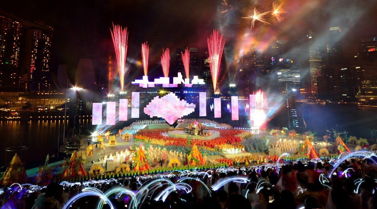 The image captures a large stage during a Chingay Parade performance, characterized by bright and colorful elements, including fireworks. The vibrant display contrasts with the dark cityscape in the background. 