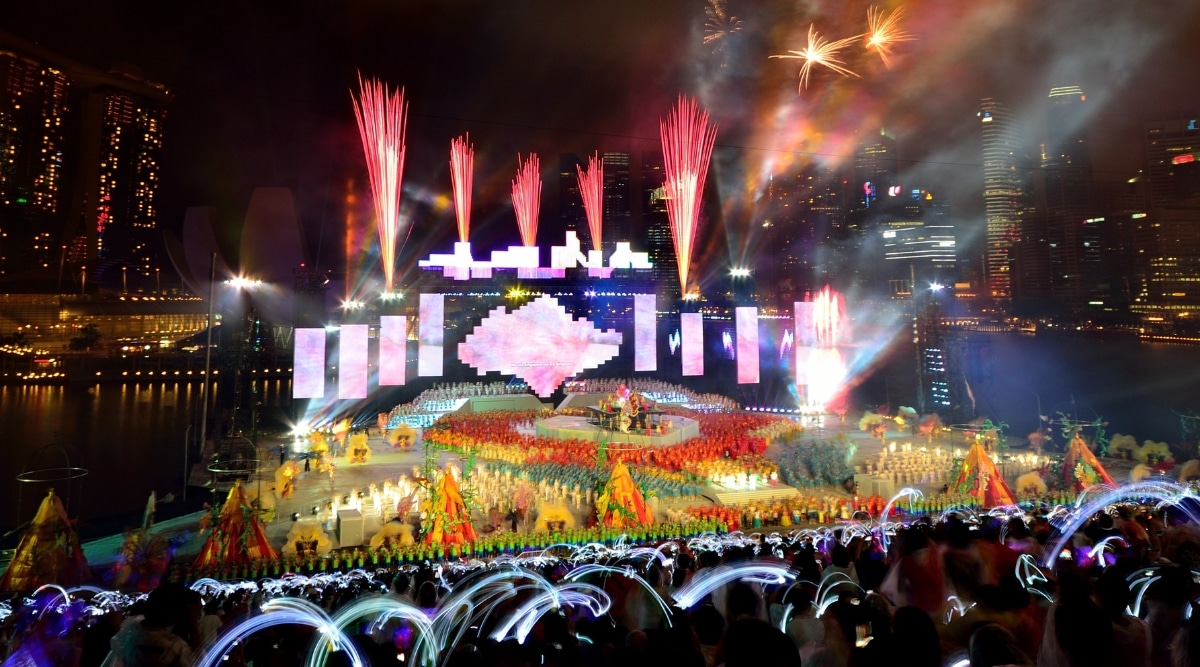 The image captures a large stage during a Chingay Parade performance, characterized by bright and colorful elements, including fireworks. The vibrant display contrasts with the dark cityscape in the background.
